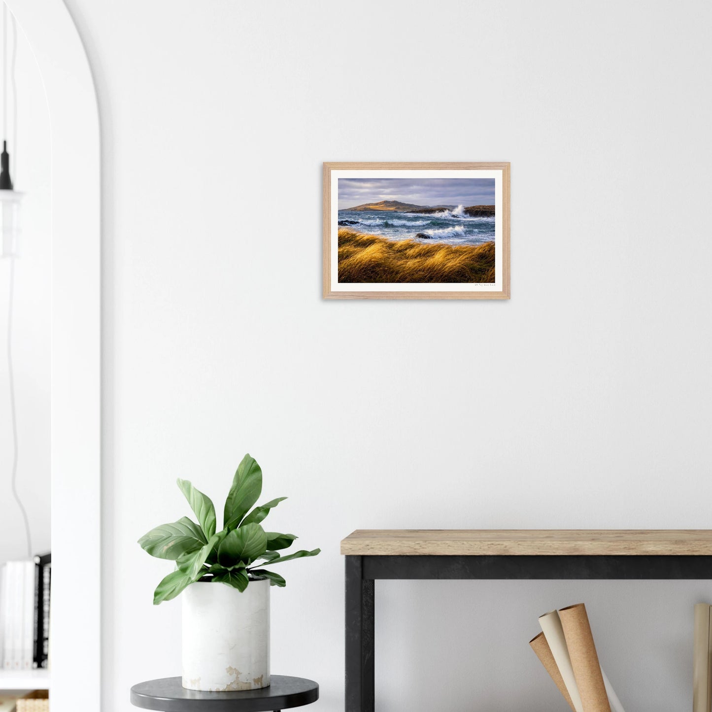 Framed artwork of a coastal scene on a white wall above a plant and table.
