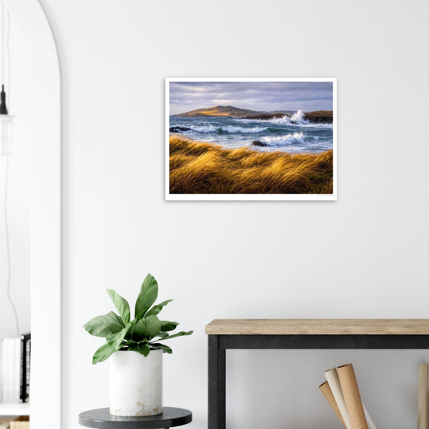 Framed landscape print of a coastal scene with waves and grass on a white wall above a desk and plant.