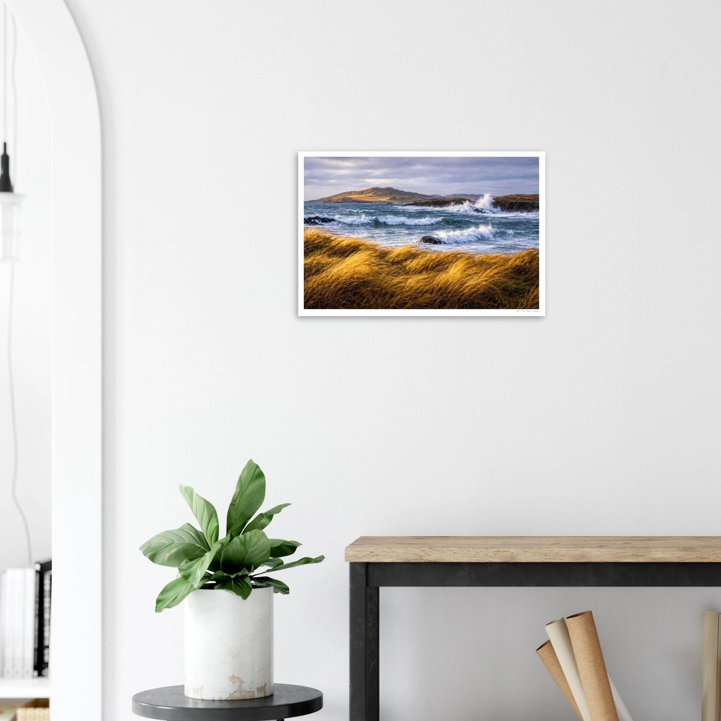 Framed artwork of a coastal scene with waves and grass on a white wall above a desk.