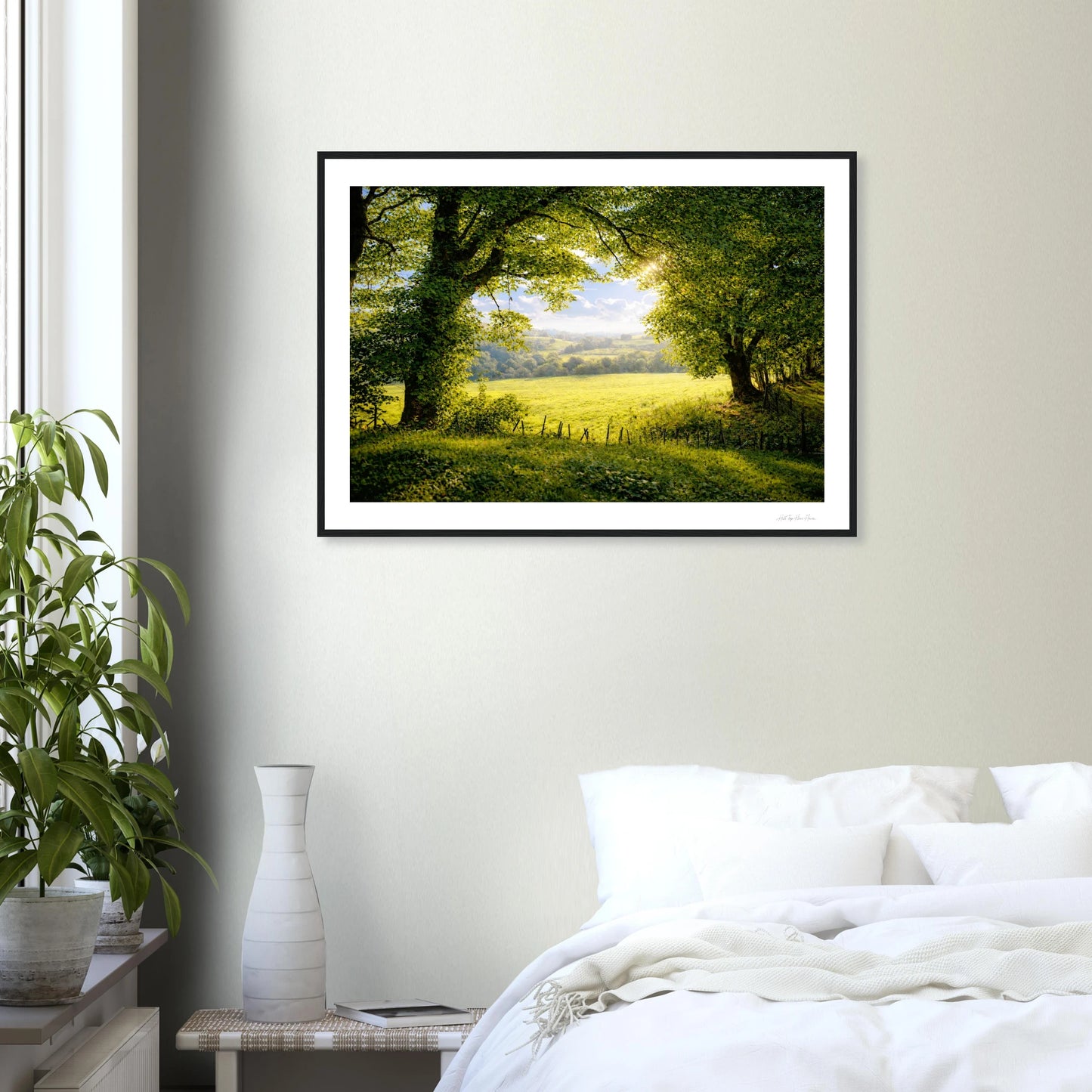 Framed landscape print of a green field with trees on a wall above a bed.