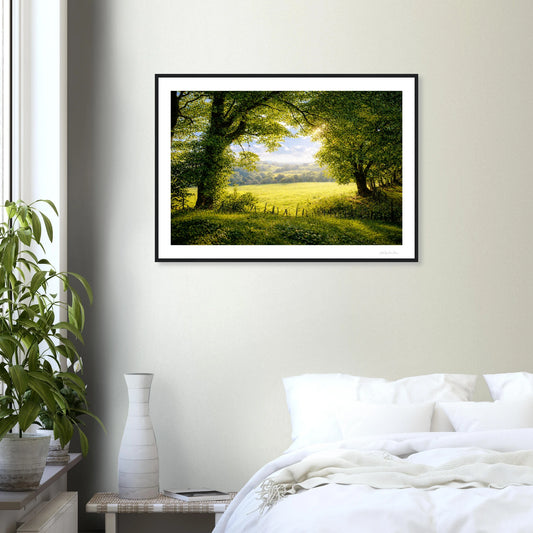 Framed landscape print of a green field with trees on a wall above a bed.