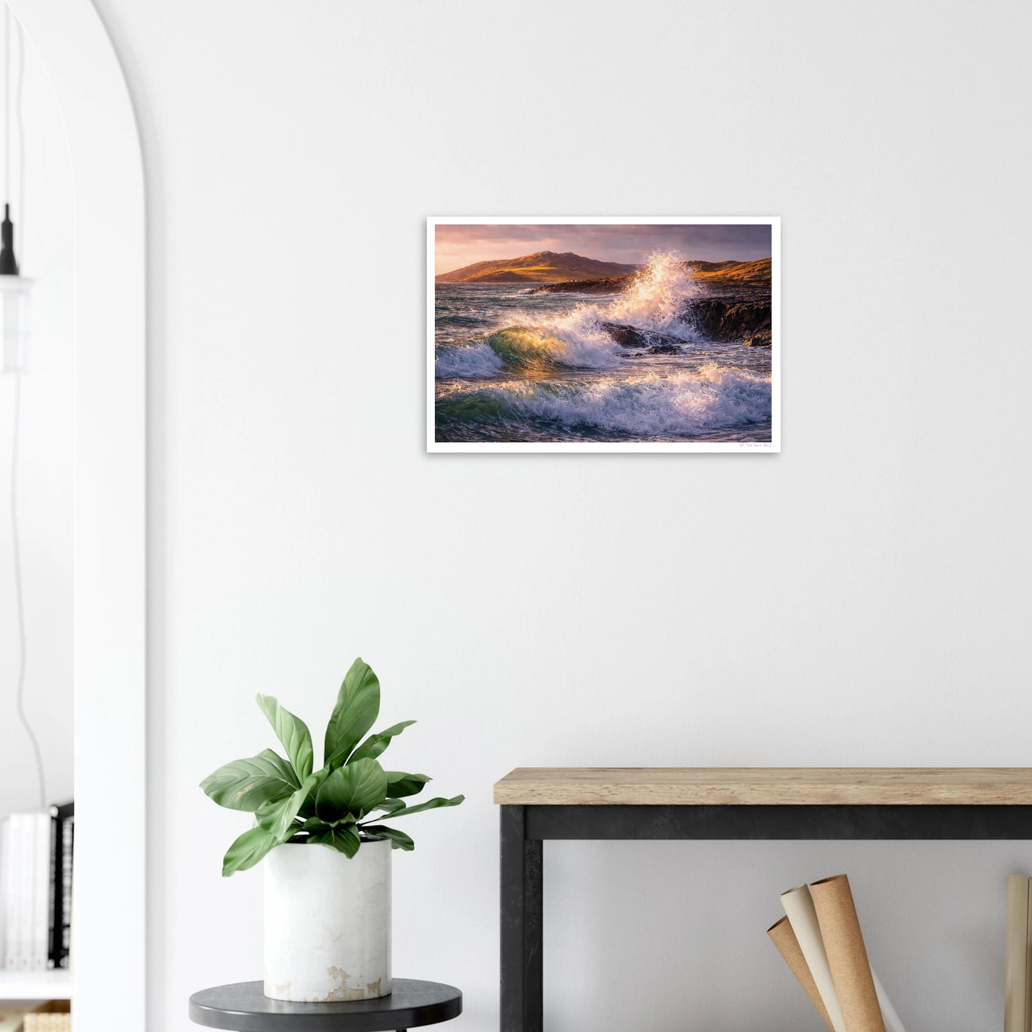 Framed artwork of a coastal scene with waves on a white wall above a desk and plant.