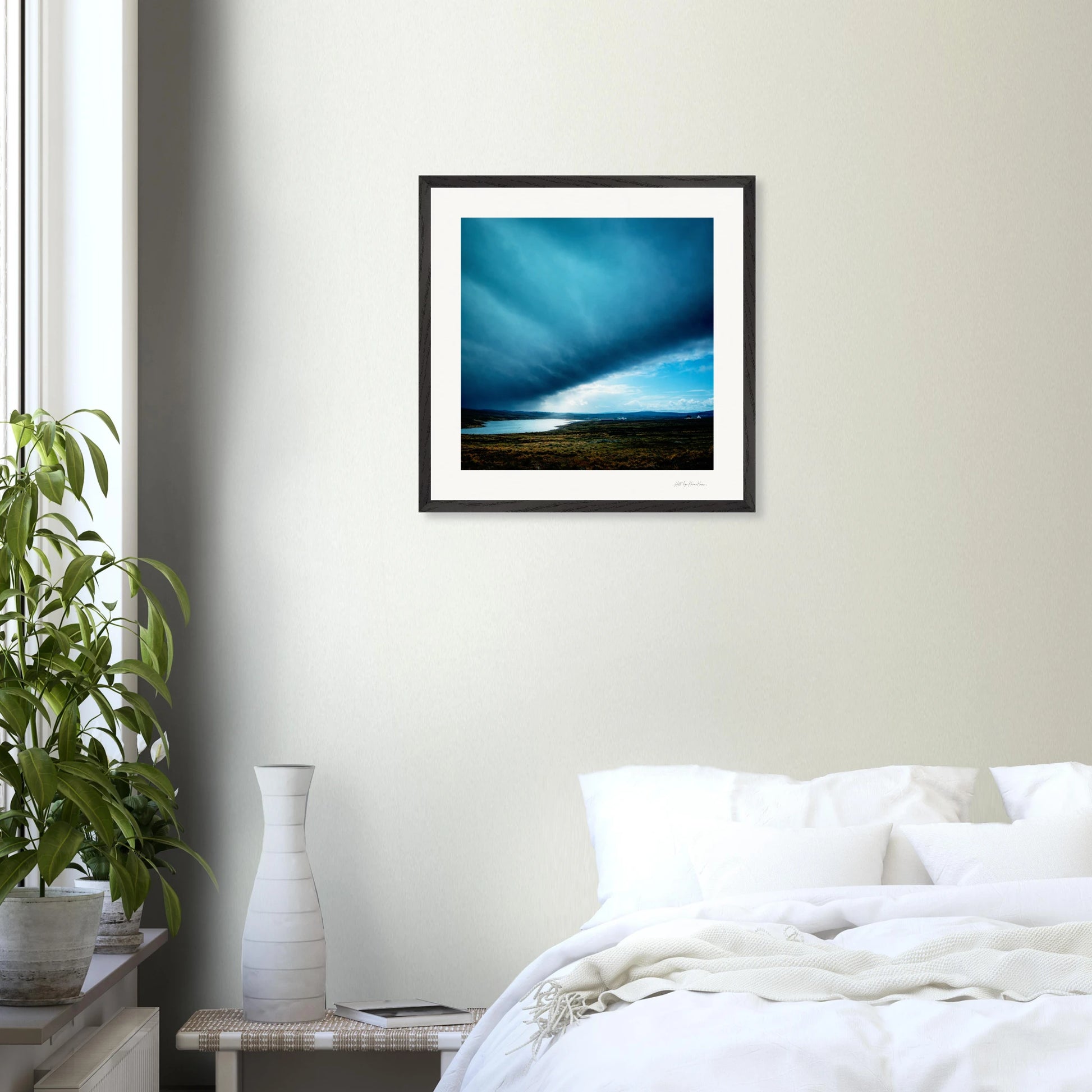 Framed artwork above a bed in a bedroom with a plant and side table.
