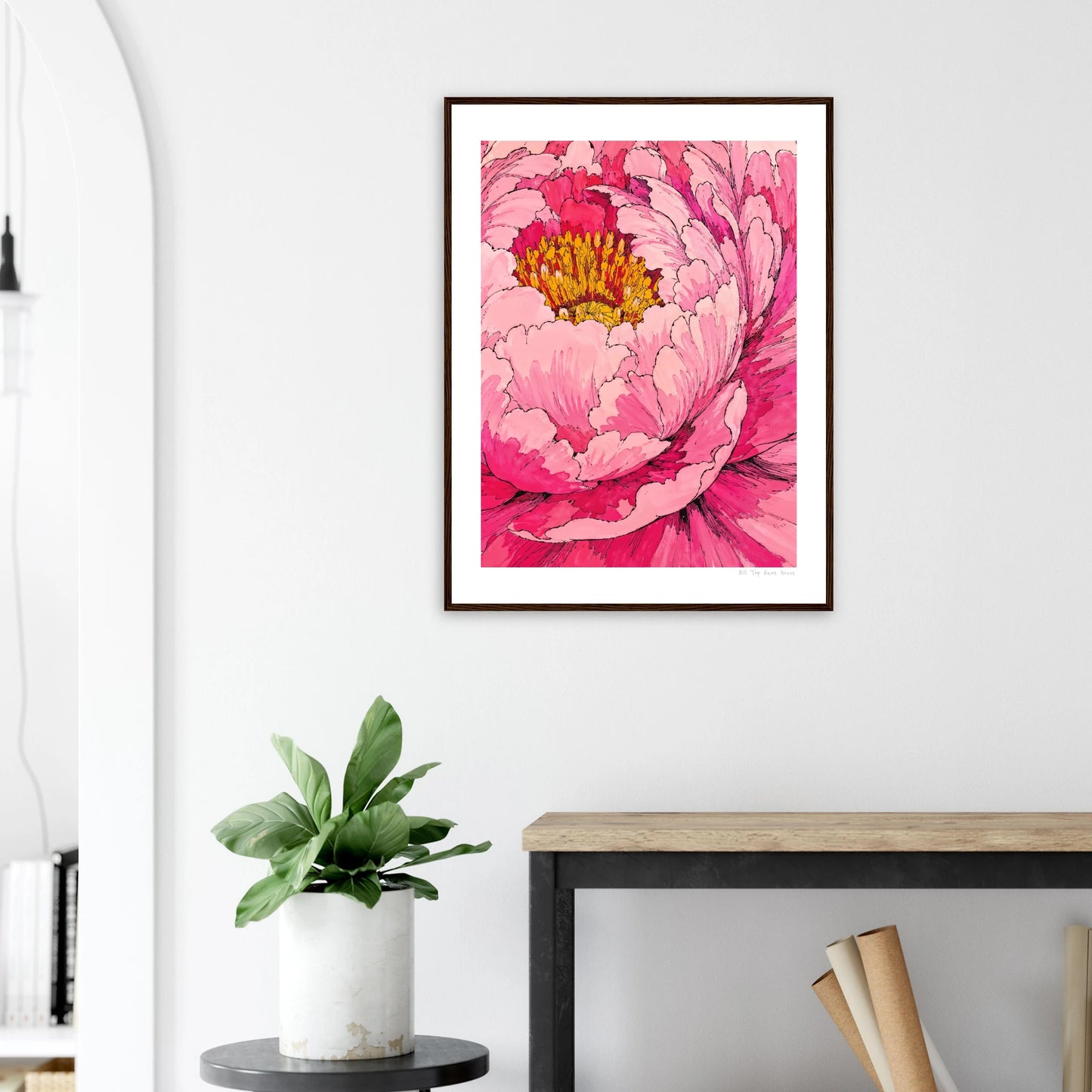 Framed pink floral artwork on a white wall above a wooden desk with a plant.