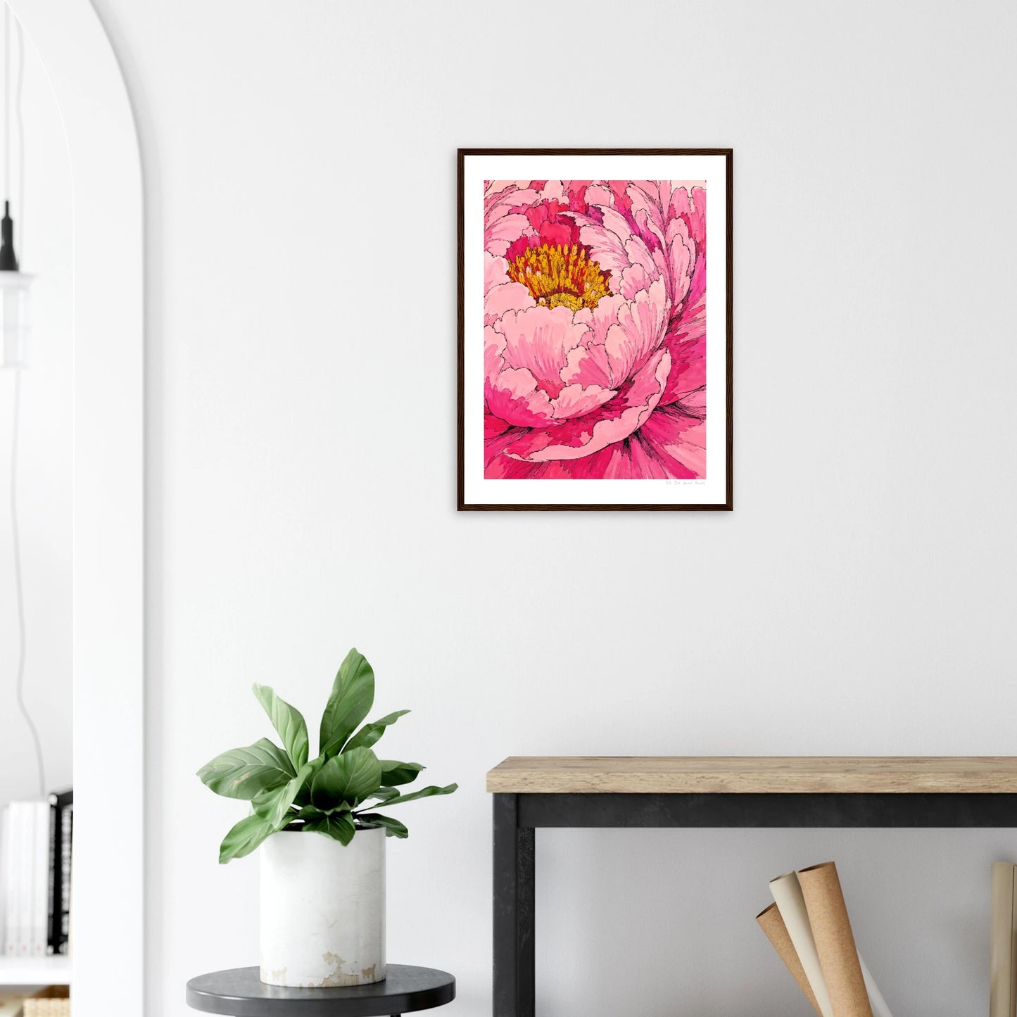 Framed pink floral artwork on a white wall above a wooden desk with a plant.