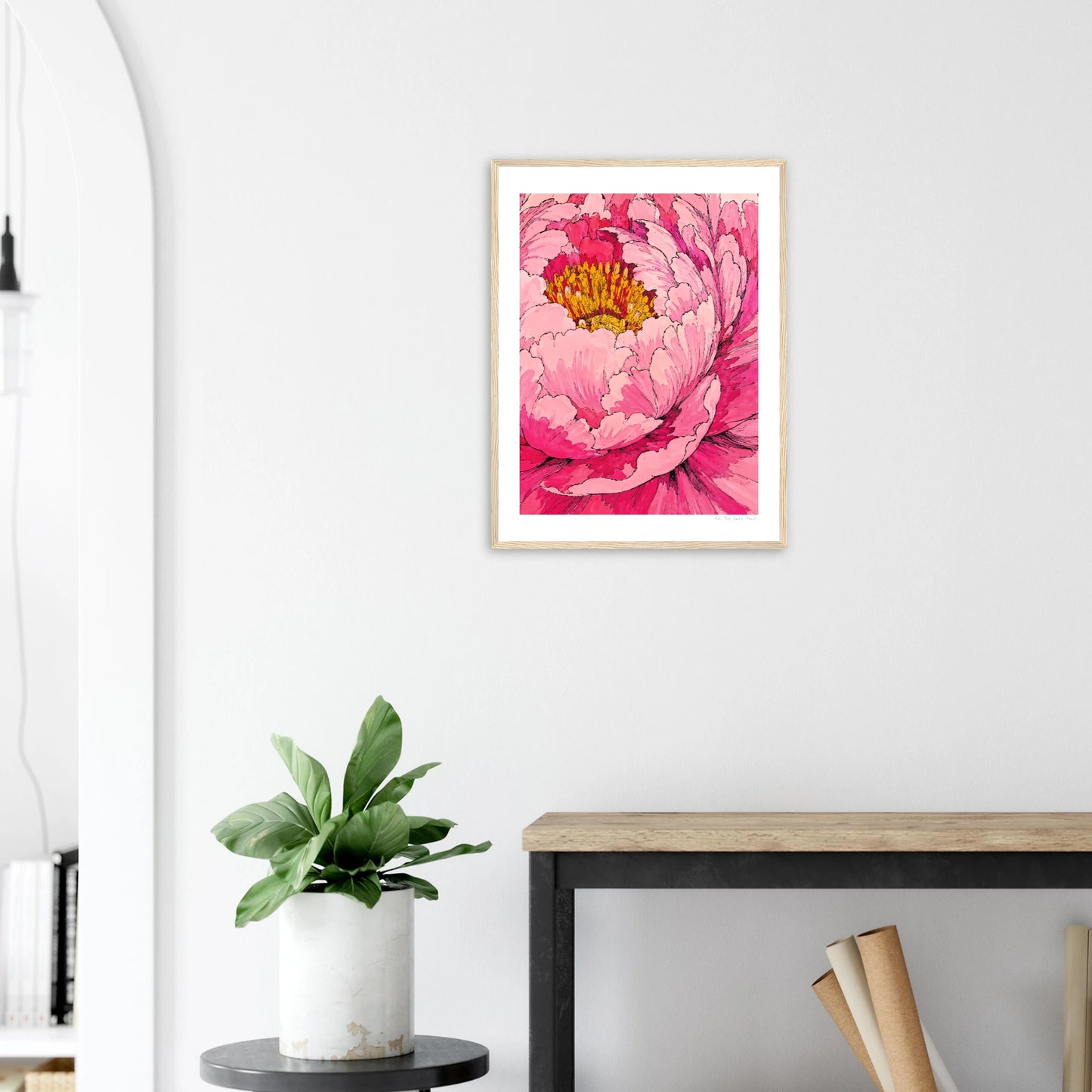 Framed pink floral artwork on a white wall above a wooden desk with a plant.