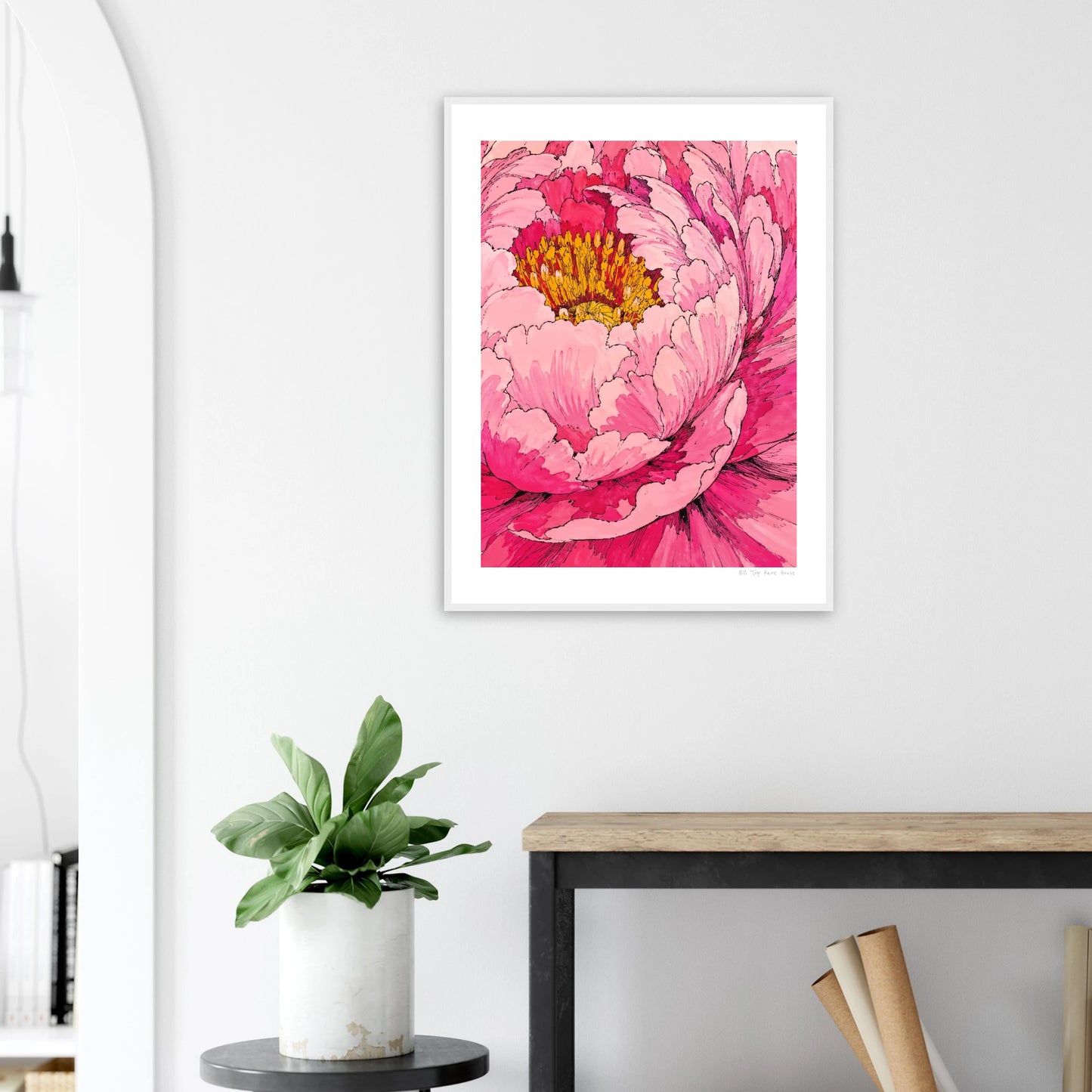 Framed pink floral artwork on a wall above a desk with a plant.