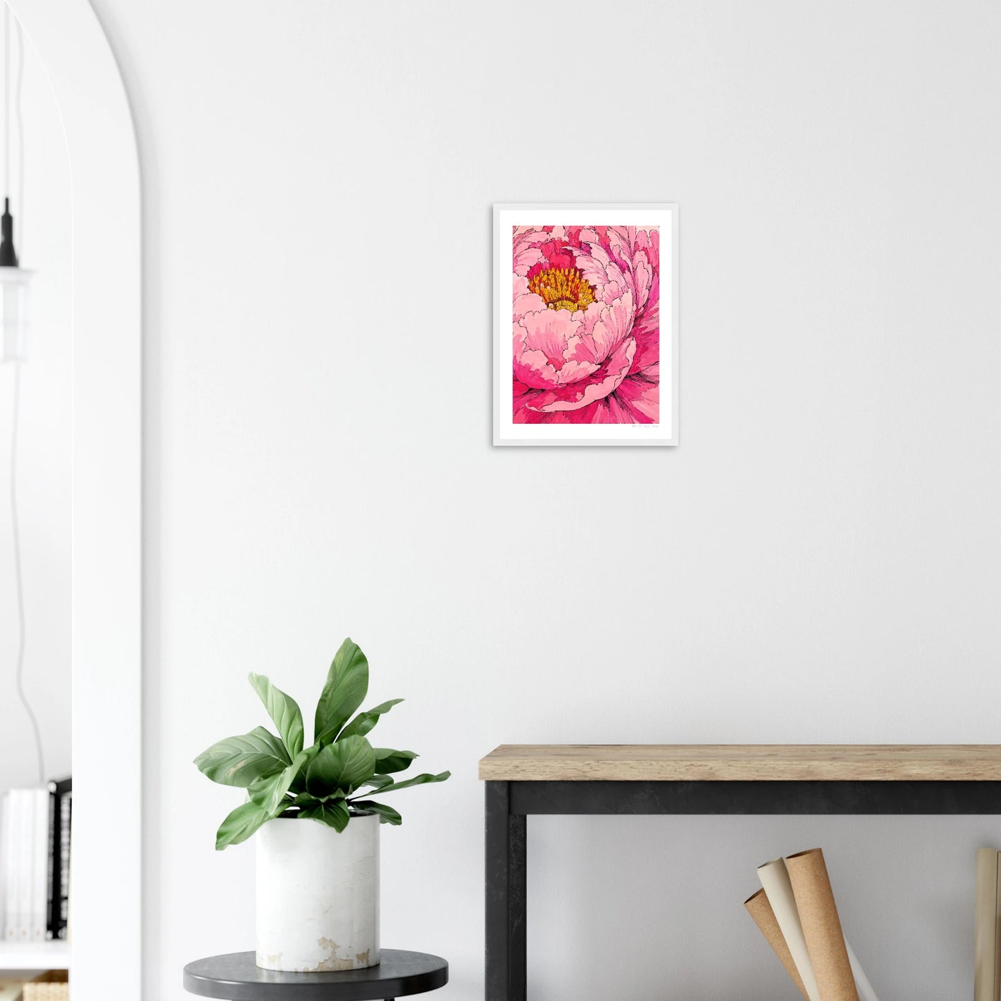 Framed pink floral artwork on a white wall above a wooden desk with a plant.