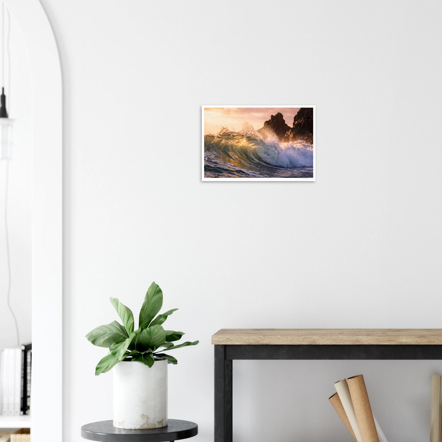 Framed artwork of a sunset over water hanging on a white wall above a plant and desk.