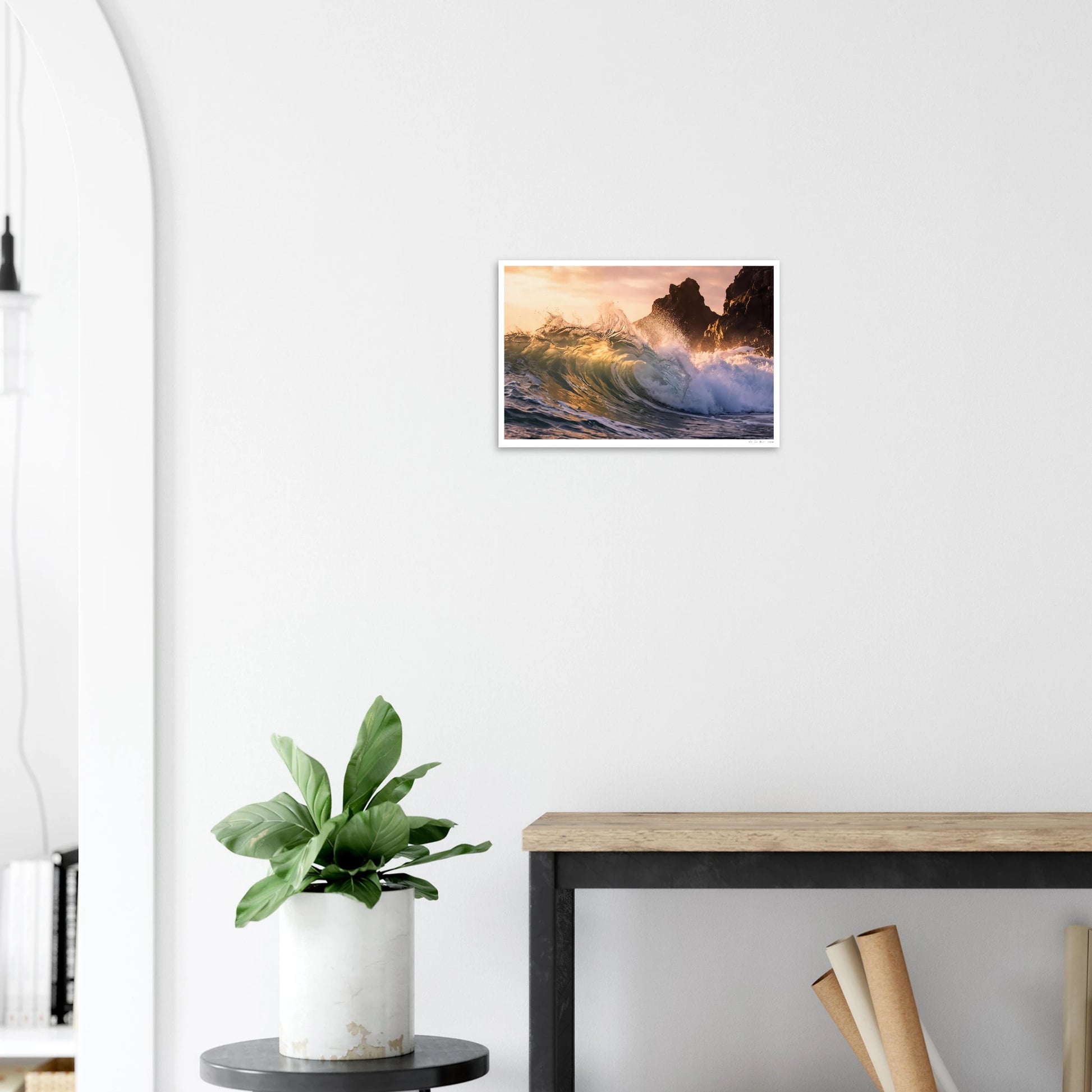 Framed artwork of a sunset over water hanging on a white wall above a plant and desk.