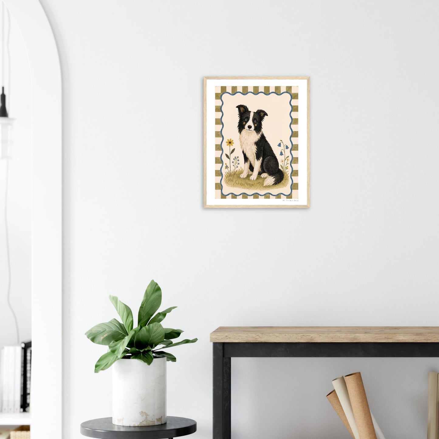 Framed artwork of a black and white dog on a white wall above a wooden desk with a plant.