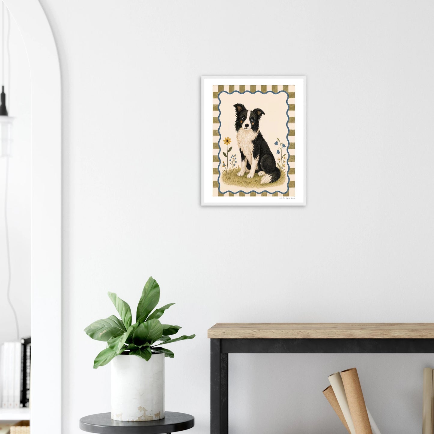 Framed artwork of a black and white dog on a white wall above a wooden desk with a plant.