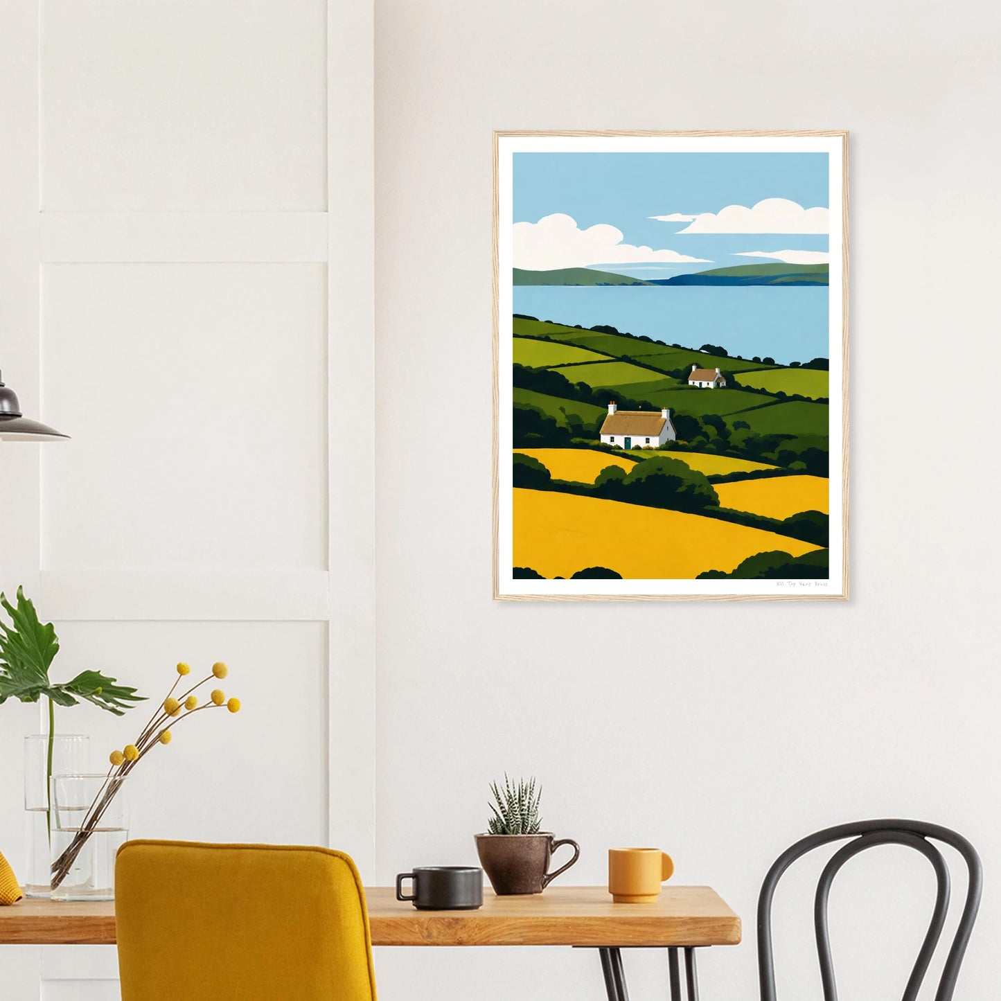 Framed artwork of a landscape with a house on a hill, displayed in a room with a table and chairs.