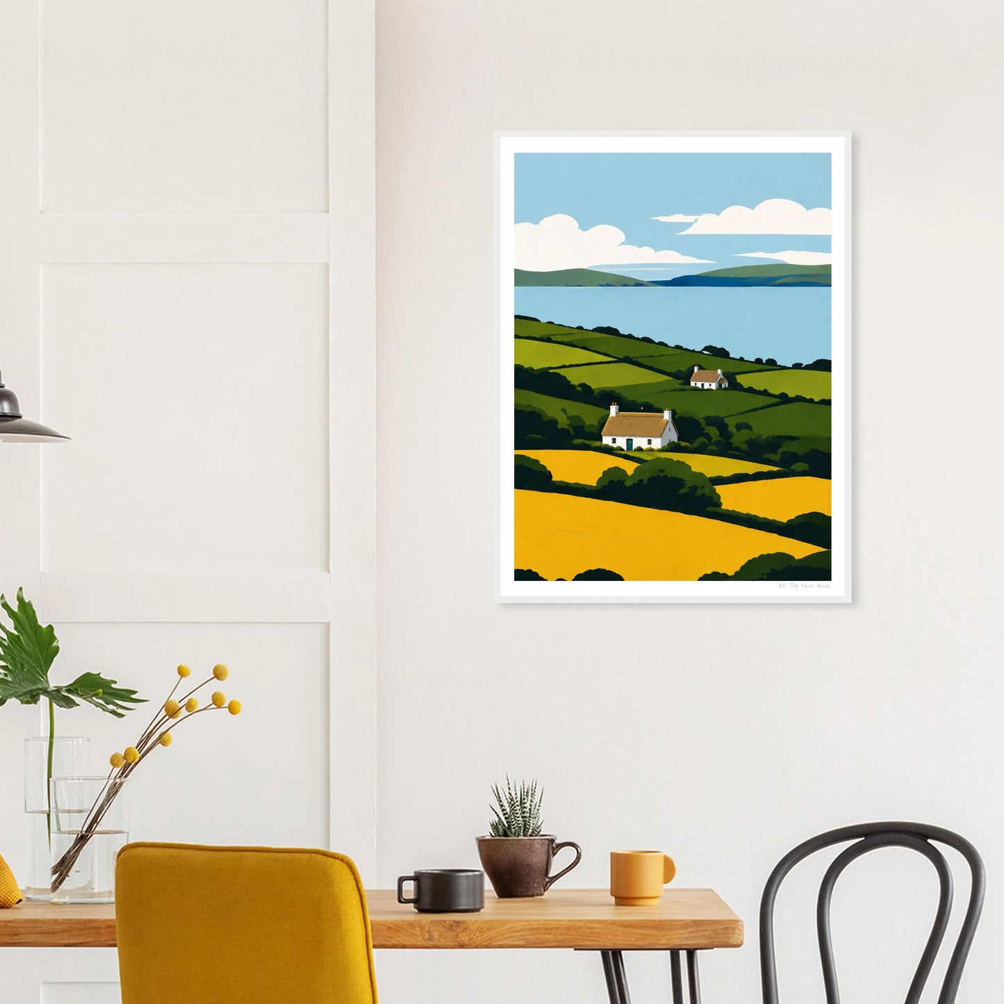 Framed artwork of a landscape with a house and fields on a wall above a table.