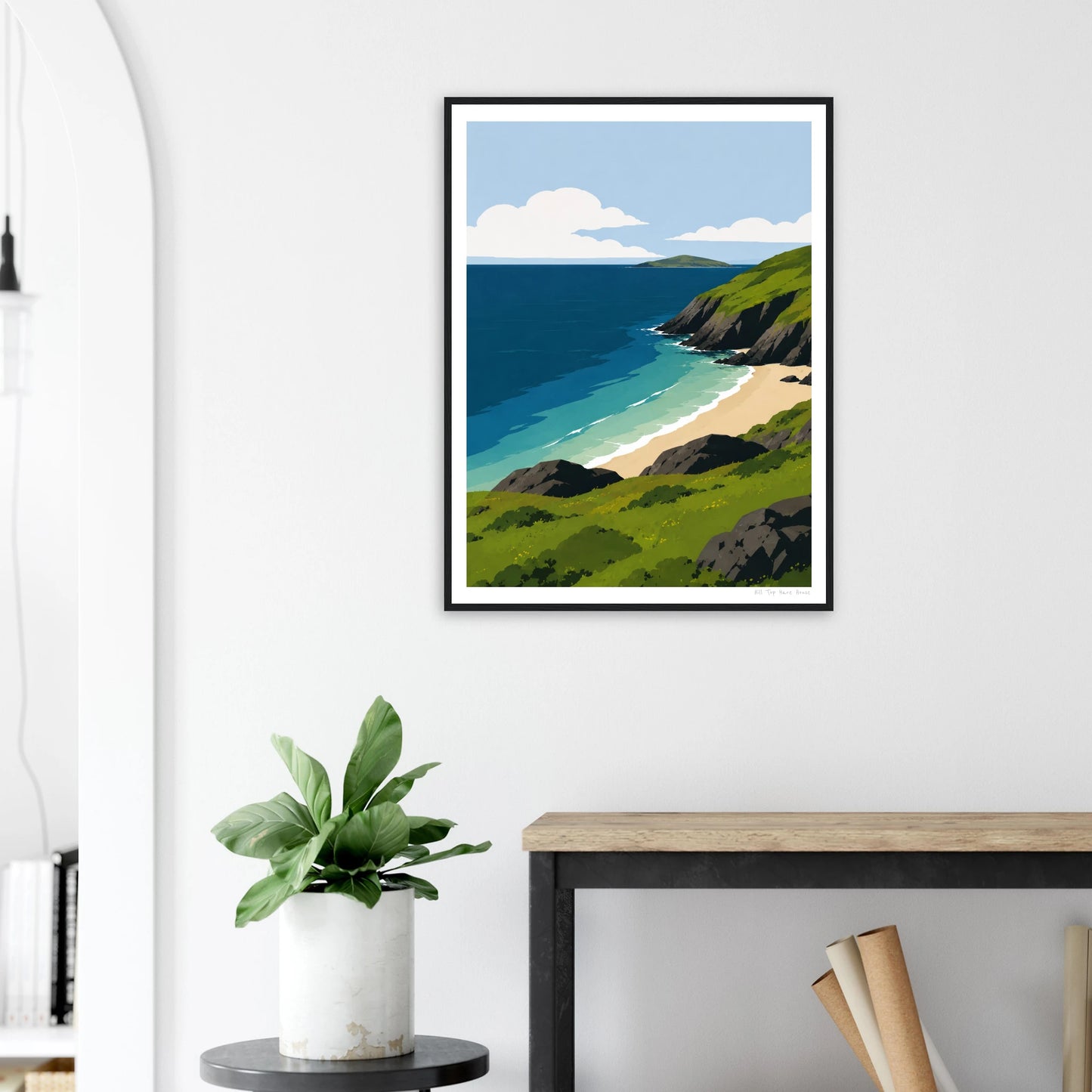 Framed artwork of a coastal scene on a white wall above a desk with a plant.
