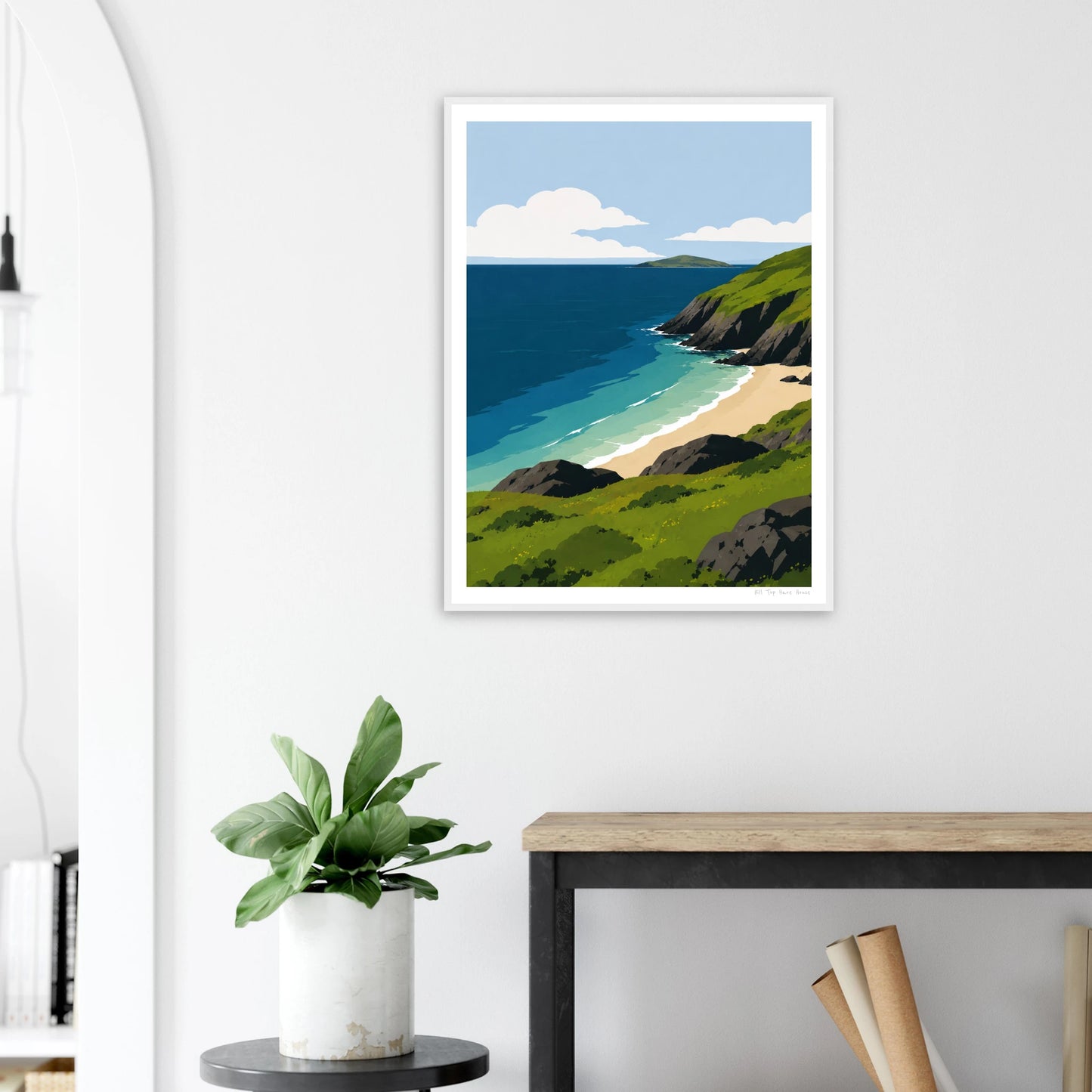 Framed artwork of a coastal scene on a white wall above a desk with a plant.
