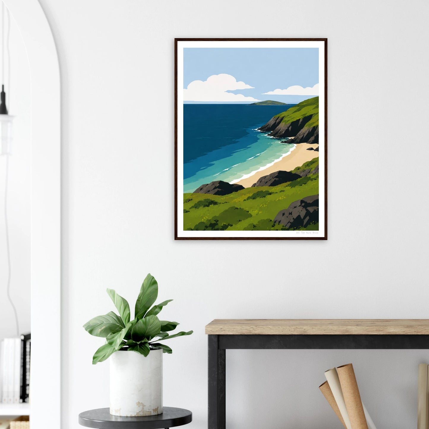 Framed artwork of a coastal scene on a white wall above a desk with a plant.