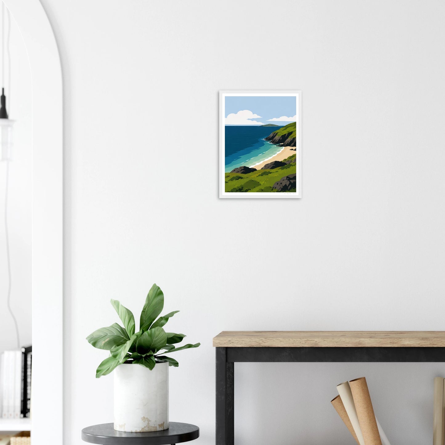 Framed artwork of a coastal scene on a white wall above a plant and desk.