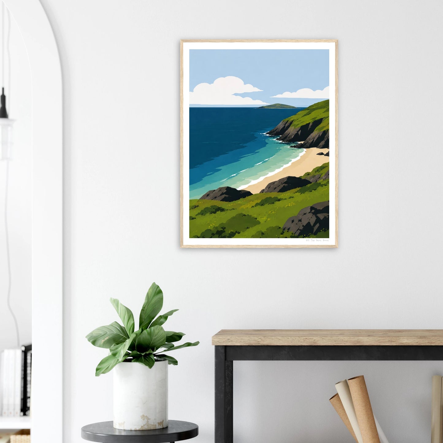 Framed artwork of a coastal scene on a white wall above a desk with a plant.