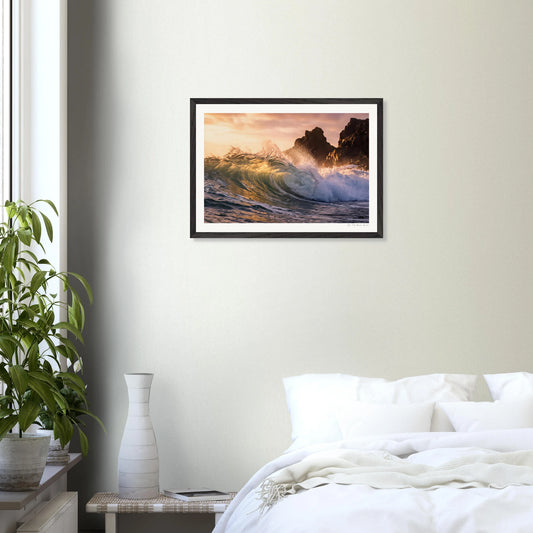 Framed artwork of a sunset over waves on a wall above a bed.